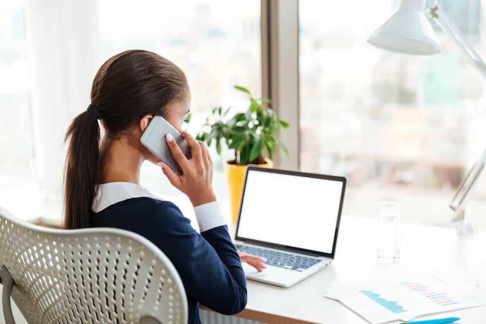 Virtual Assistant with blue cardigan and white shirt sitting in front of a computer while she speaks on the phone. In the table there are some documents a blue pen, a glass of water a plant with yellow base, and a light bulb. She is in a room with big windows.