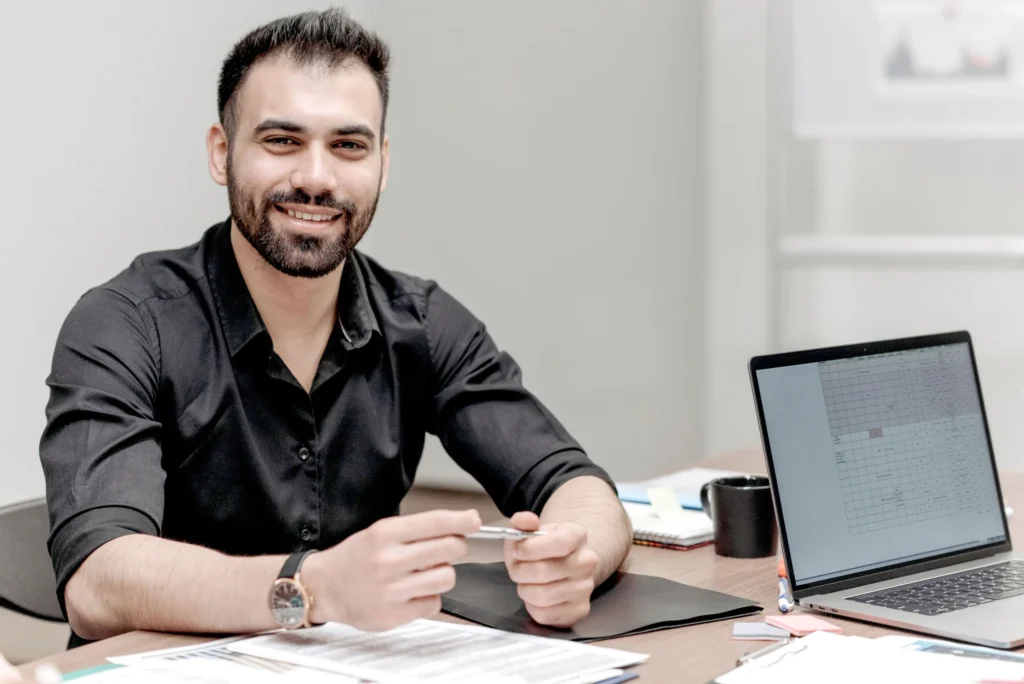 smiling man wearing a black shirt and a watch, holding a pen with both hands while sitting at a desk that has folders, notebooks, notepads and a laptop on top
